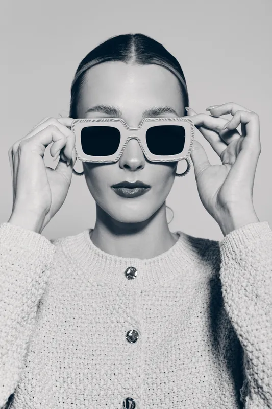 Portrait of a young white woman with light skin wearing sunglasses and a knit sweater, posing confidently in studio lighting.