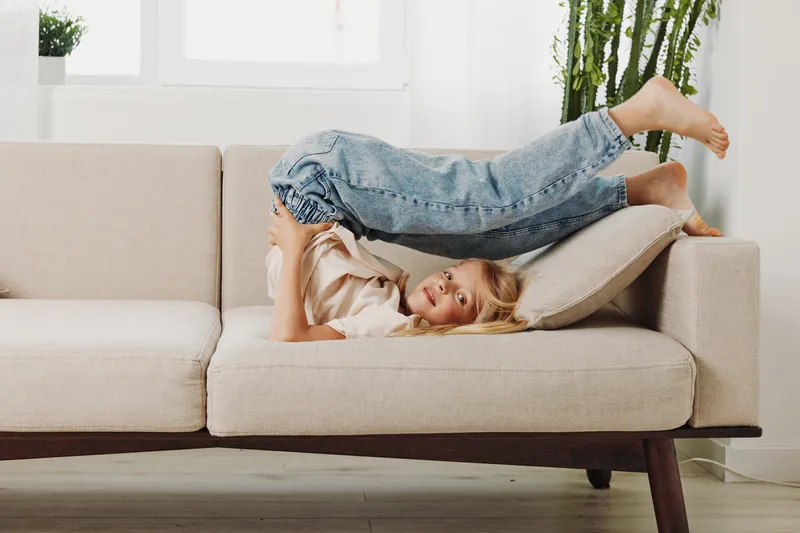 Playful child in casual clothes lies upside down on beige sofa with cushion in bright living room. Concept of childhood joy, home comfort, and relaxed lifestyle.