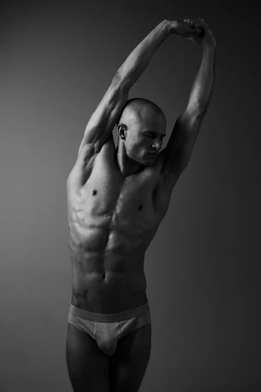 Black and white photo of a muscular man flexing, showcasing strength and fitness. The dramatic lighting highlights the contours of his body in an artistic way.