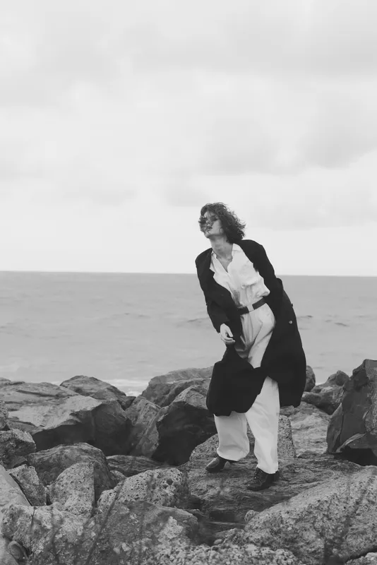 Woman standing on jagged rocks by the sea in a long coat, windy weather, monochrome fashion with a dramatic pose in an outdoor seascape