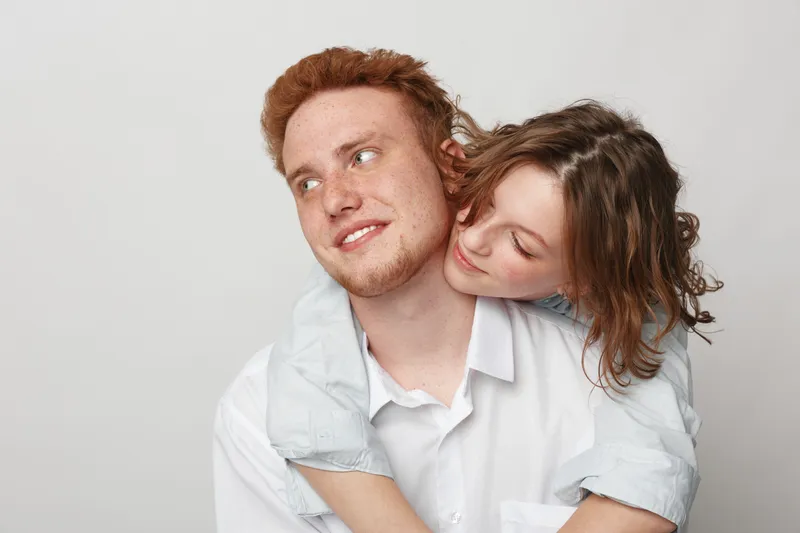 Young couple in casual clothes share affectionate moment, woman hugs man from behind with smile. Intimate relationship, love, happiness, and connection on neutral background.
