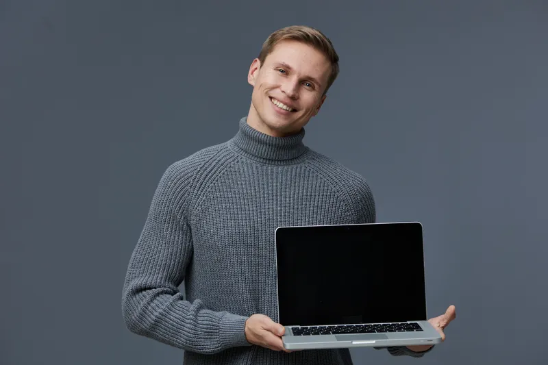 Smiling man wearing gray turtleneck sweater holding open laptop computer isolated on gray background studio portrait. Technology, casual fashion, cheerful expression, modern device, professional, rela