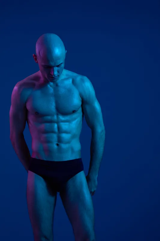 Athletic man posing with dramatic blue lighting, showcasing his toned physique and expressing confidence in a captivating and artistic manner.