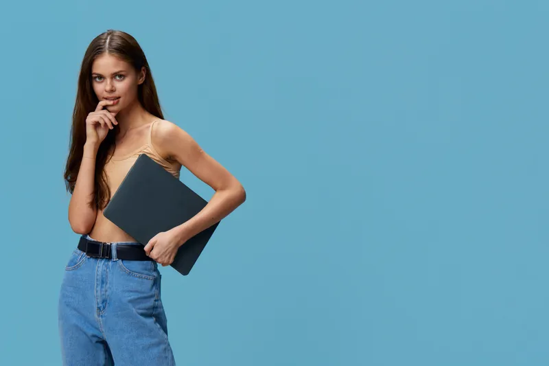 young woman holding laptop with thoughtful expression, wearing casual beige top and blue jeans against solid blue background, modern tech vibe