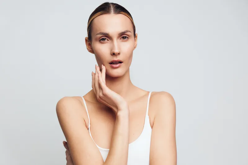 Young slender woman with natural makeup and slicked hair poses with hand on face showing clean skin and calm expression isolated on white background in studio.