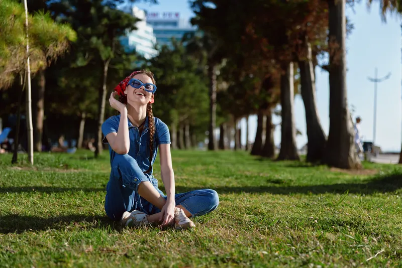 lifestyle woman in modern boho-western denim street style sitting on grass outdoors with film color effect, sunglasses, casual summer vibe, sunny day