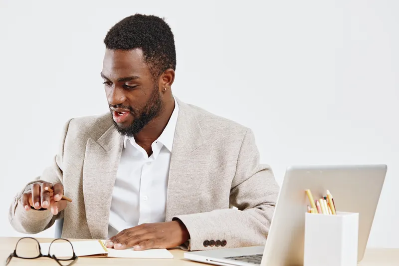 Professional young African American man working at modern office desk with laptop, notebook, and glasses on white background. Business, technology, workplace, education, productivity, professional con