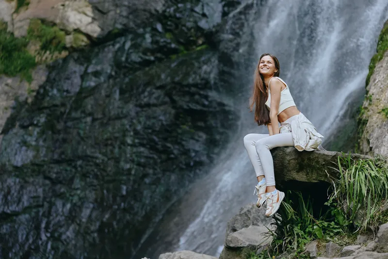 smiling woman sitting near waterfall in casual white outfit, surrounded by rocks and green plants, enjoying outdoor nature adventure and fresh air