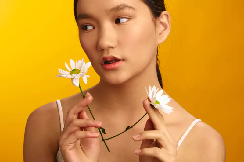 asian woman gazes softly at the camera, holding white daisies against a bold yellow backdrop, a studio portrait that conveys warmth, grace, and a contemplative mood.