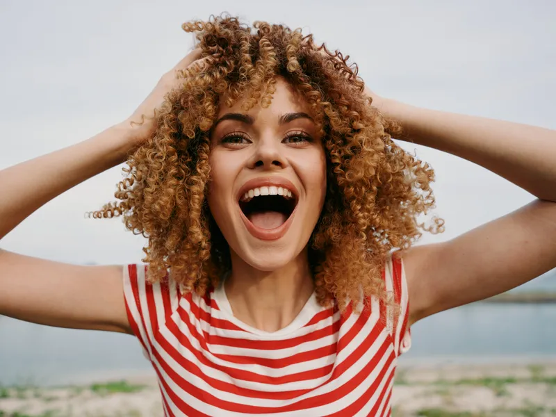 A joyful woman with curly hair wearing a red and white striped shirt stands on a sunny beach, expressing exuberant happiness with an open smile and lively energy.