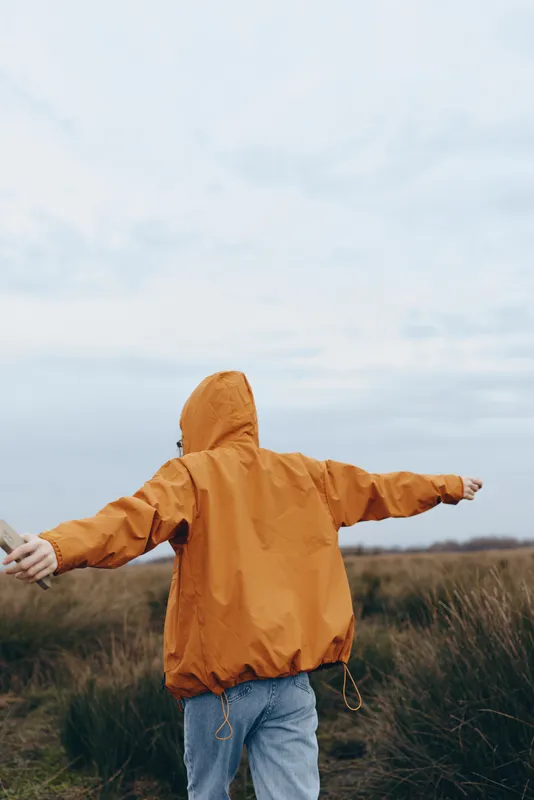 Woman lifestyle outdoors, raincoat hood up, arms out in grassy field, orange jacket and jeans, facing cloudy sky, casual fashion, calm mood