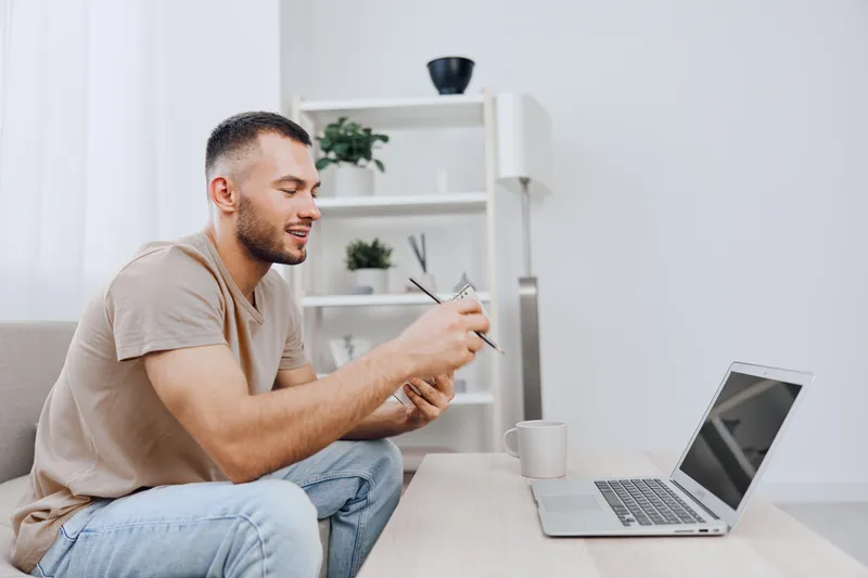 Young man working on a laptop, smiling and focused, surrounded by a cozy home environment with plants and bright natural light. Ideal for remote work themes.