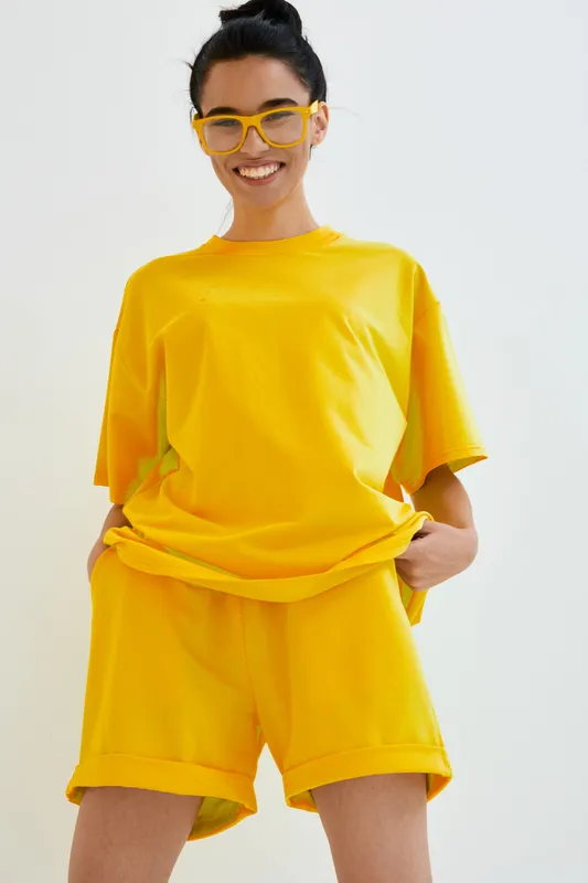 smiling woman in bright yellow casual outfit and matching glasses stands confidently against white background, showing joyful expression and relaxed posture