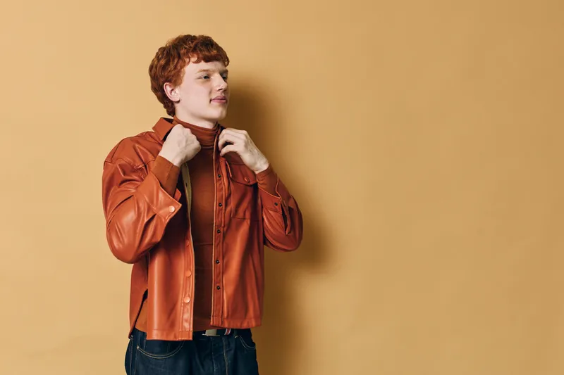 Young man with red hair wearing stylish orange jacket and turtleneck sweater posing confidently against beige background in casual fashion portrait.