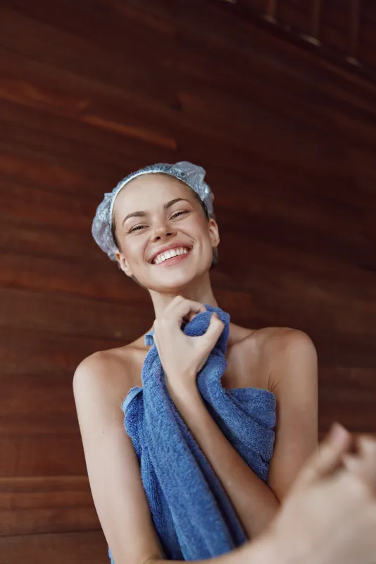 Smiling woman wrapped in blue towel with shower cap showing happiness and freshness against dark wooden wall background, natural skincare, beauty routine, wellness concept.