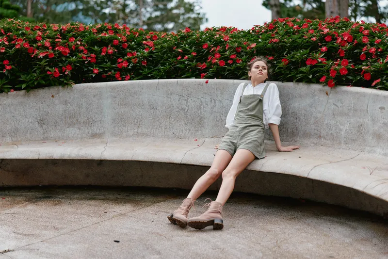 Young woman in casual green overalls, relaxing on a curved stone bench surrounded by vibrant red flowers, exuding a carefree attitude in a serene park setting