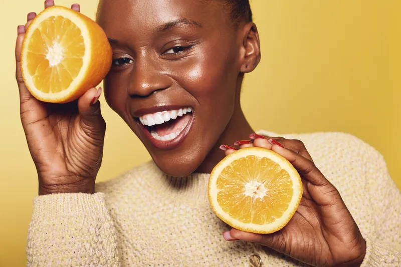 Happy dark skinned African woman in beige sweater holds two halves of fresh orange fruit near face on yellow background. Concept of healthy lifestyle, vitamin C, wellness and natural beauty.