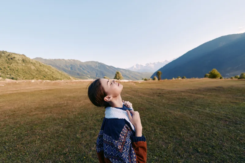 Woman in sweater portrait standing in a wide mountain meadow outdoors, eyes closed and breathing deeply, enjoying nature and scenic landscape in a peaceful, calm travel moment.