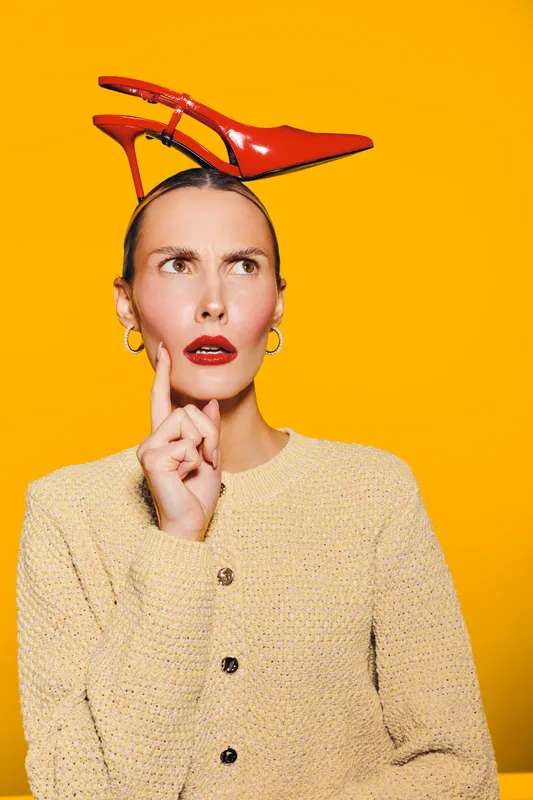 Thoughtful woman with red high heel shoe on head looks puzzled, wearing beige sweater and gold earrings. Bright yellow background creates vibrant contrast and playful mood.