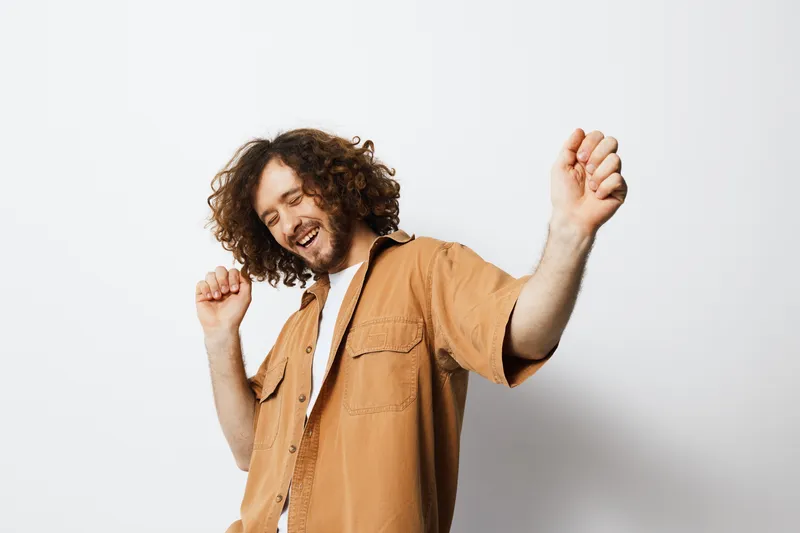 Cheerful young man dancing joyfully in a light brown shirt against a white background, expressing happiness and carefree emotions in a vibrant, energetic scene