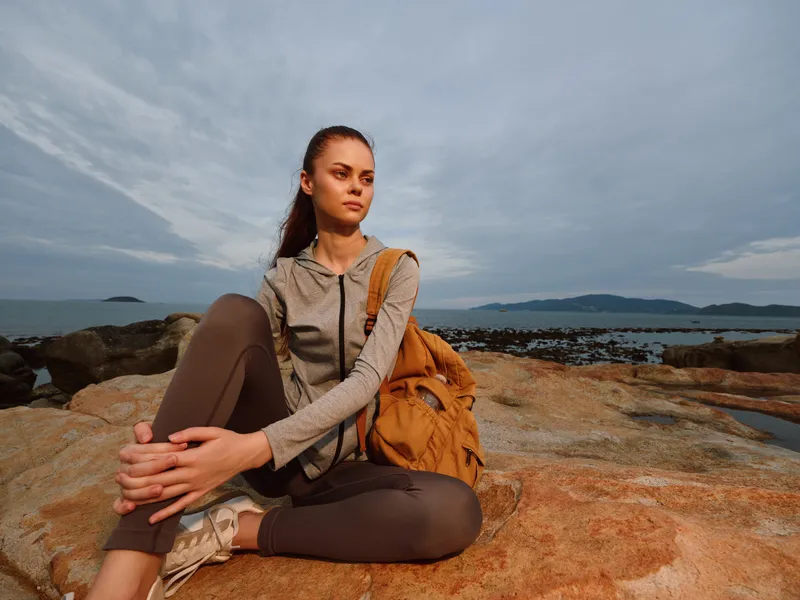 Young woman in activewear sitting on rocks by the sea, showcasing a thoughtful expression against a cloudy sky, ideal for wellness and outdoor lifestyle themes