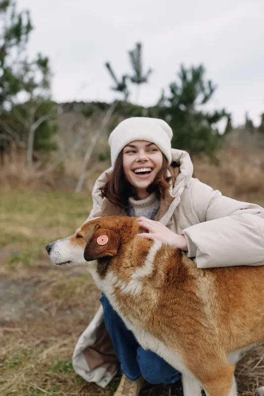 Woman in a warm coat and beanie hugs a large dog in an open outdoor field, smiling widely as they share joyful companionship. Soft natural light and a grassy landscape create a cozy, candid moment of