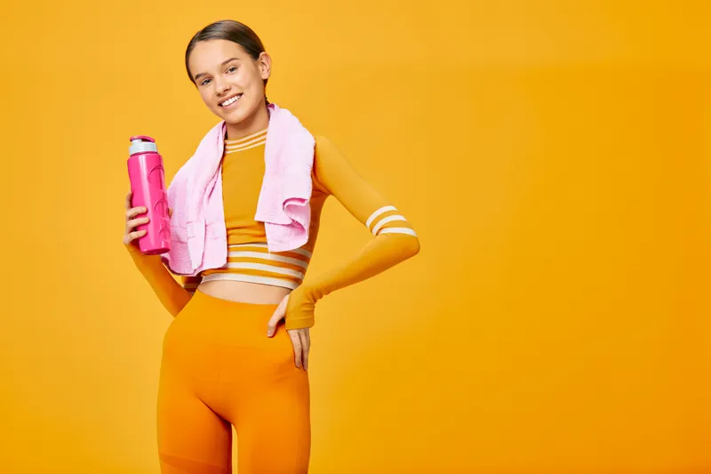 Woman in bright orange athletic outfit with a towel over her shoulder and a water bottle, posed against a vivid orange background, showcasing fitness, sport and vibrant healthy living.