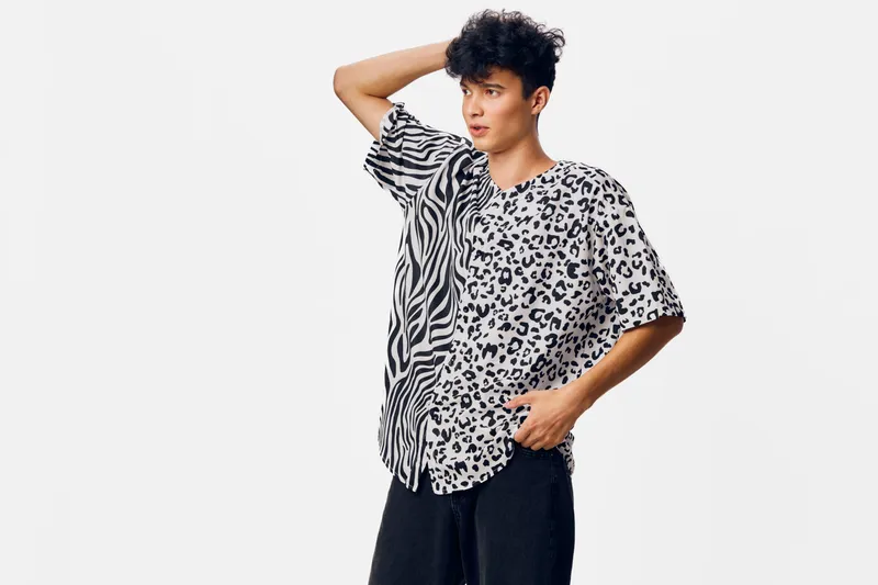 Young man wearing a black and white patterned shirt with zebra and leopard prints, posing with one hand behind his head against a plain white background.