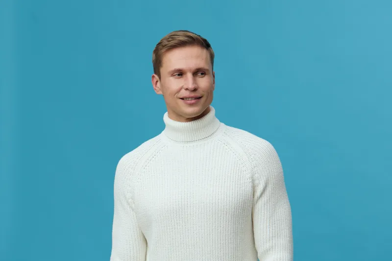 Smiling man in white sweater with short brown hair posed against blue background studio portrait. Casual, happy, cheerful, male, lifestyle, fashion, relaxed.