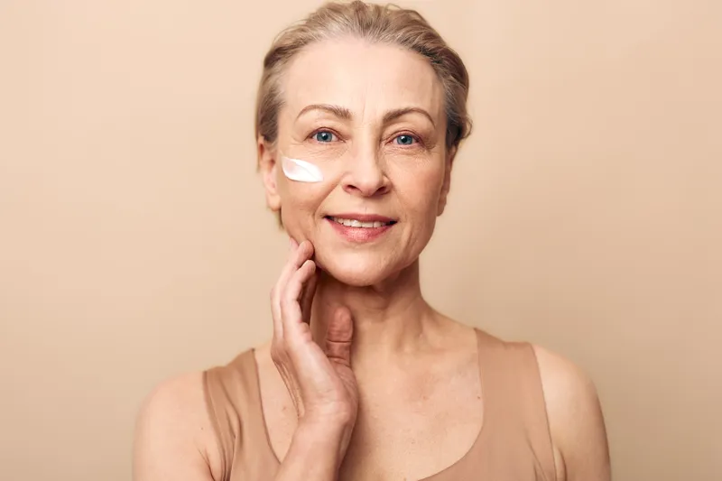 Mature woman with natural skin applying face cream on cheek, smiling gently against beige background. Concept of skincare, aging beauty, self-care and healthy skin routine for older adults.