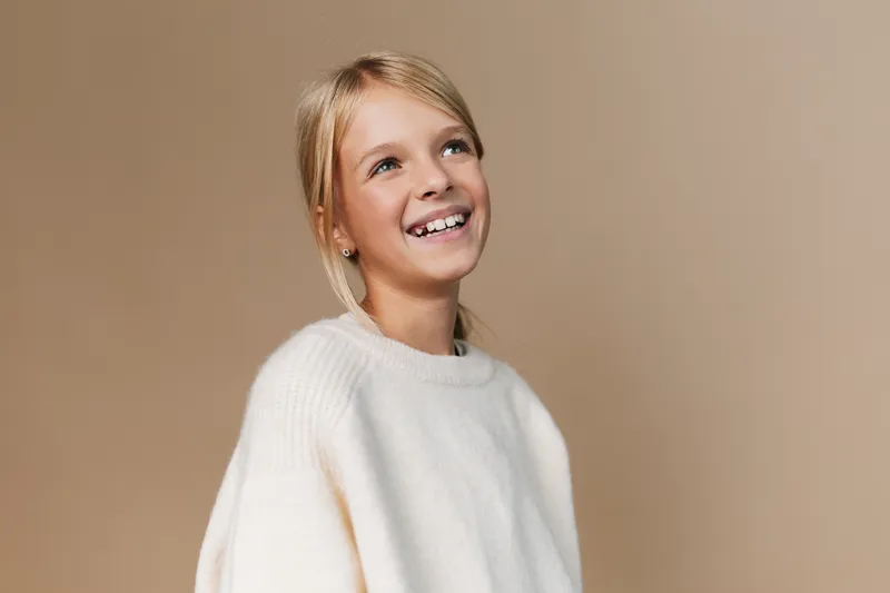 Authentic portrayal of a joyful school age girl smiling in a cozy sweater, capturing everyday play and routine moments with genuine charm and warmth.