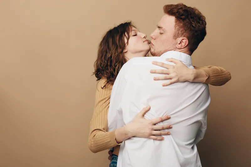 Young couple embraces and kisses passionately on beige background. Woman in brown sweater hugs man in white shirt. Concept of love, romance, intimacy, and affection between partners.
