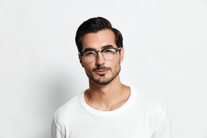 Stylish young man with glasses poses confidently while looking at the camera, showcasing his casual white t-shirt against a minimalistic background.