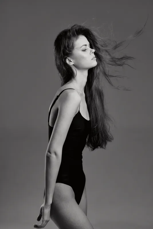 Black and white portrait of a slender woman in a black bodysuit with flowing hair against a gray backdrop