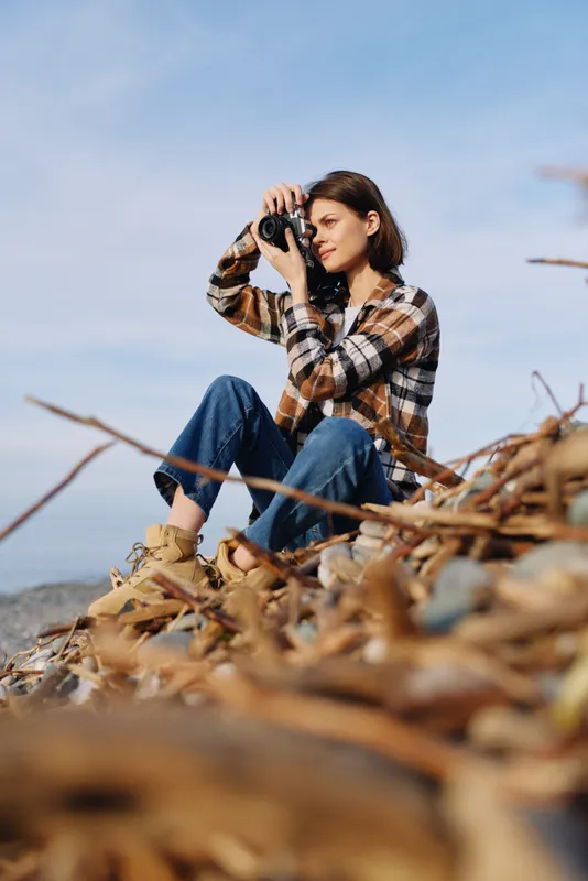 Photographer woman crouching among autumn leaves on a rocky shore, eyes at the viewfinder, capturing landscape and light in daytime outdoor scenery.
