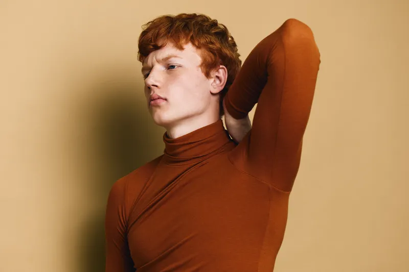 Redhead man in brown turtleneck posing with hand behind neck against beige background, portrait with thoughtful expression and casual fashion style for studio photography.