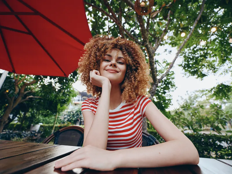Main focus on a young woman with curly hair seated at a wooden table under a red umbrella, her gentle smile framed by soft daylight and lush outdoor greenery.