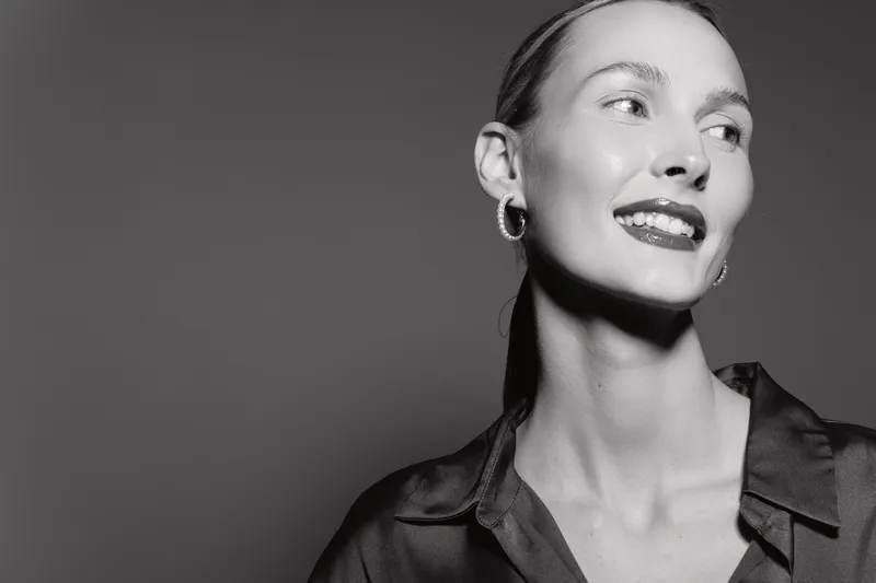 Portrait of a smiling Caucasian woman with fair skin wearing earrings and dark blouse against a gray background in monochrome studio lighting.