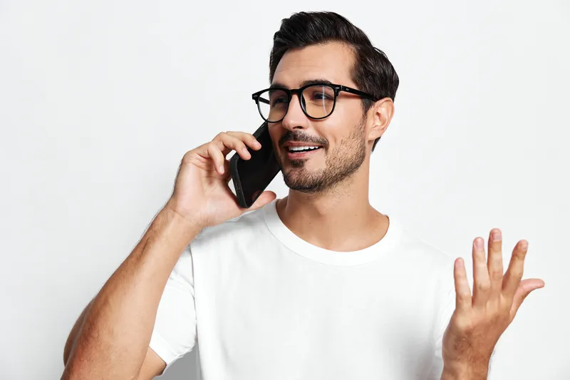 Smiling man wearing white t-shirt talking on mobile phone, expressing joy with hand gestures, isolated on plain light background. Communication lifestyle concept.
