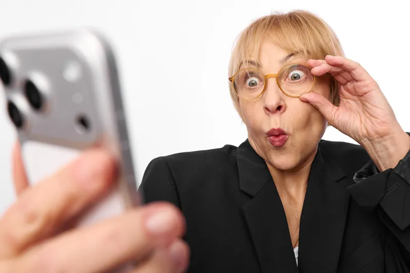 Surprised woman in office, holding glasses near face and touching hair while looking at smartphone, wide eyed shock expression, soft indoor light, blurred phone