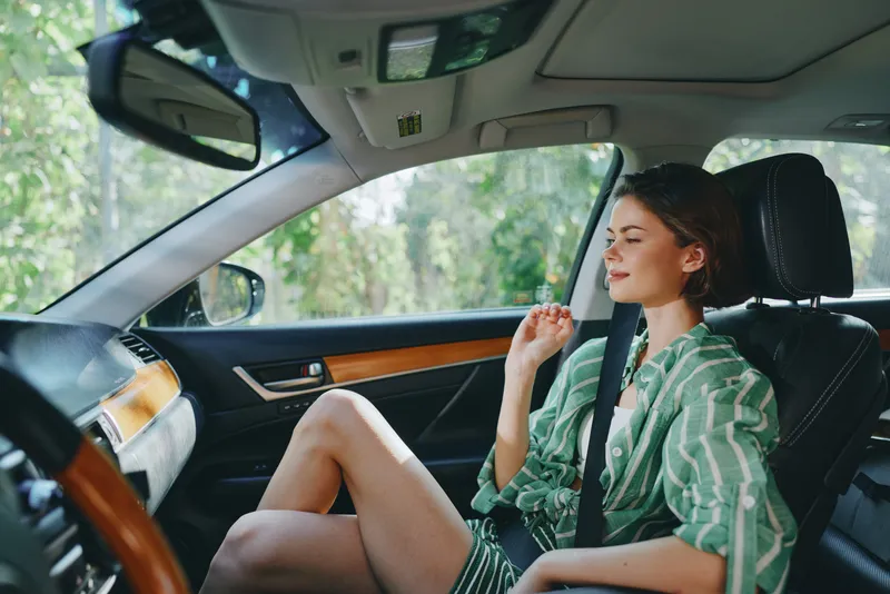 Young woman sits in a car seat with a seatbelt on, relaxed pose, soft daylight, and striped shirt, emphasizing style and comfort.