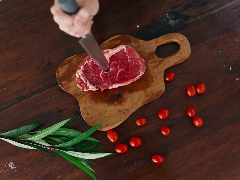 Женщина с ножом в руке нарезает свежее стейковое мясо для жарки на кухне; на столе соль, перец и другие специи, красные помидоры черри и зелень, приготовление ужина