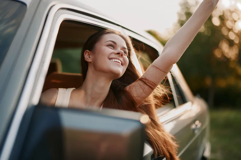 Young woman enjoying a road trip, smiling with her arm out of the car window, highlighting freedom and joy in nature during sunset.