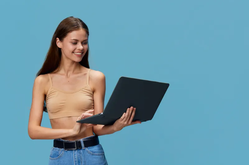Young woman holding laptop with smile, wearing casual beige top and blue jeans, standing against plain blue background, showing technology use and positive mood.