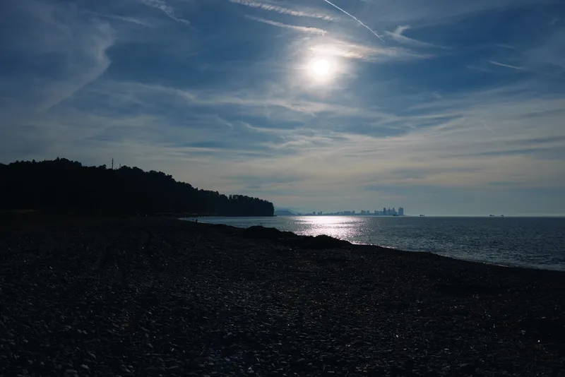 Beach scene with a pebbled shore, calm ocean, and sun reflecting off the water, a distant silhouette of land on the horizon under a blue sky, creating a tranquil coastal mood.