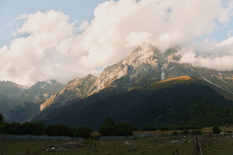 Mountain landscape with rocky peaks partially covered by clouds under blue sky. Green forest and grassy field in the foreground. Concept of nature and outdoor adventure.