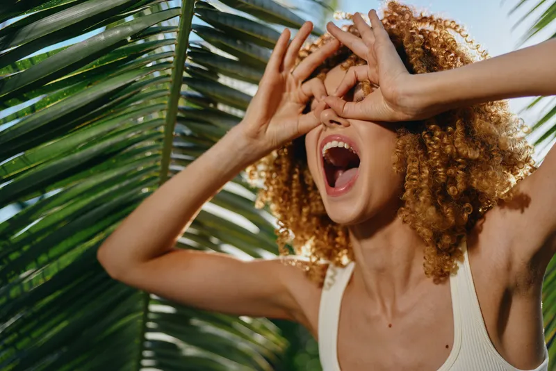 Happy woman with curly hair making binoculars gesture by tropical palm leaves in bright sunlight, expressing joy and playful mood during summer outdoors.