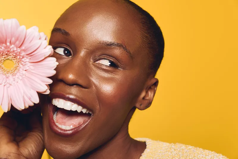Happy African woman with dark skin holds pink flower near face smiling brightly on yellow background. Concept for beauty, skincare, wellness, and natural joy with vibrant colors.