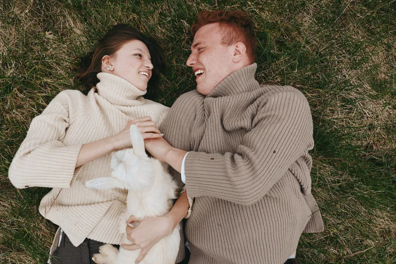 Lifestyle couple outdoors, lying on grass, smiling as they cradle a small goat kid between them, warm sweaters, soft sunlight, natural colors, candid moment, relaxed mood, rural atmosphere
