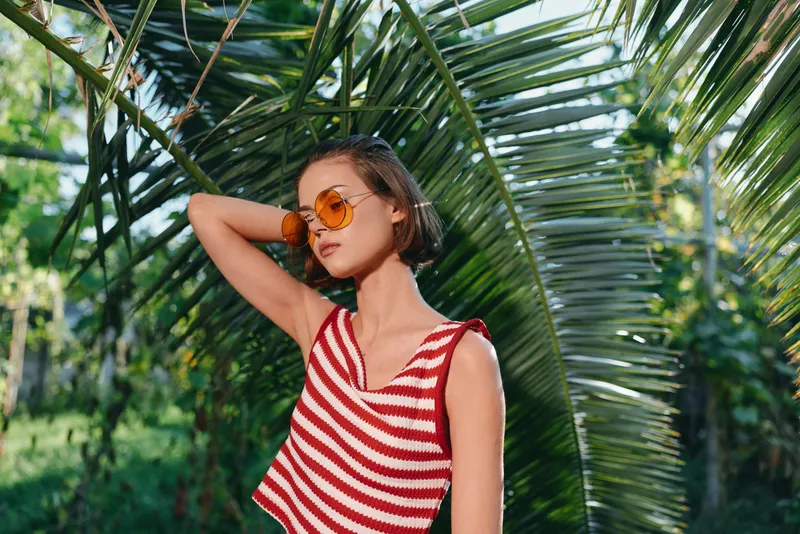 A stylish young woman poses outdoors in bright sunlight, wearing a red and white striped sleeveless top and orange sunglasses. She exudes confidence and a carefree, sunny mood amid lush greenery.